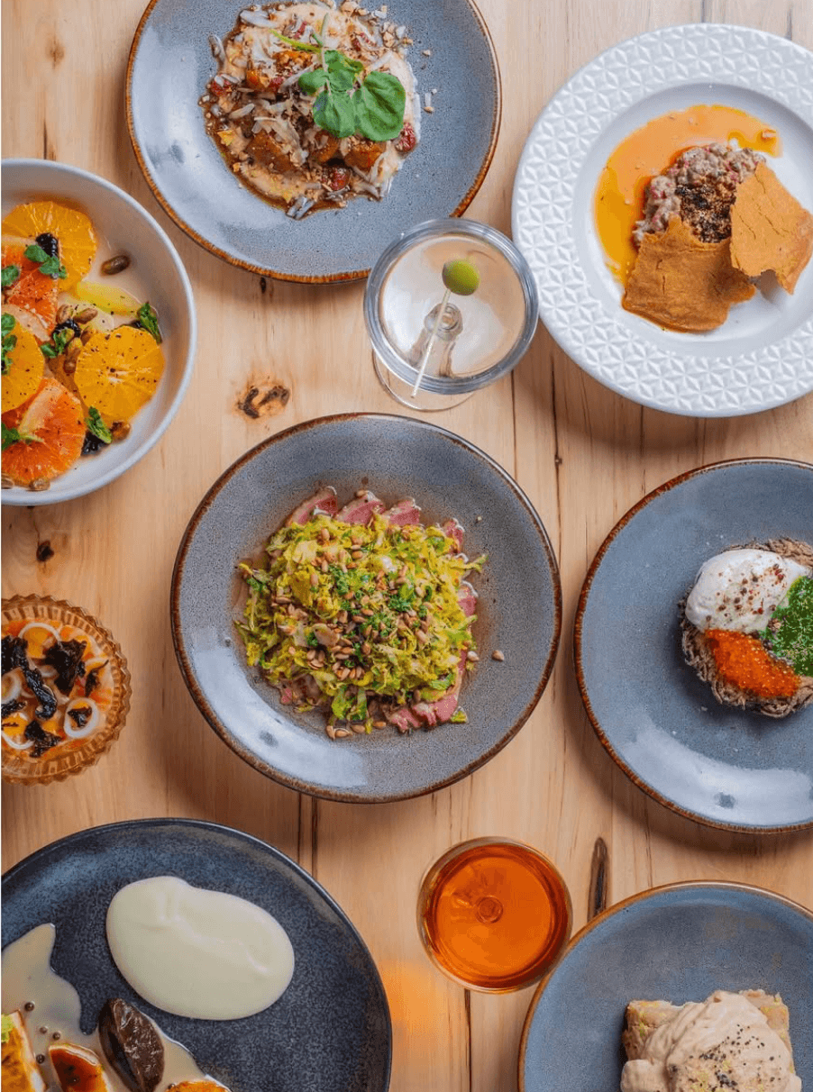 An overhead view of a wooden table in Saratoga Springs with assorted gourmet dishes, including salads, tart, meatballs, mashed potatoes, and drinks, all artfully plated on gray and white plates. Hamlet and Ghost in Saratoga